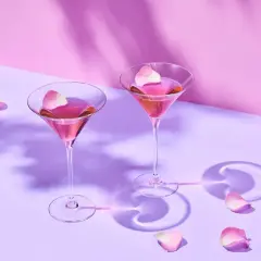 Dwa kieliszki z różowym martini, ozdobione płatkami róż. Delikatne cienie i pastelowe tło tworzą romantyczną atmosferę.