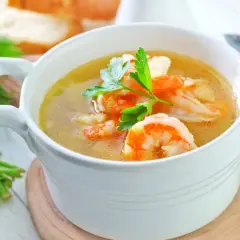 Aromatyczna zupa krewetkowa z krewetkami i natką pietruszki w białej miseczce, obok pęczek ziół i kromki chleba.
