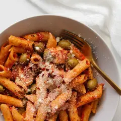 Penne z sosem pomidorowym, oliwkami i startą mozzarellą, posypane płatkami chili. Złoty widelec czeka na pierwszy kęs.
