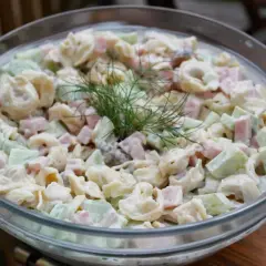 Pyszna sałatka z tortellini, szynką i ogórkiem, udekorowana gałązką koperku. Idealna na letnie przyjęcie.