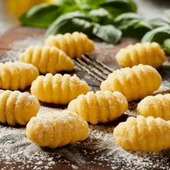 Świeżo przygotowane, puszyste makaron gnocchi, posypane mąką, leżą na drewnianej desce obok gałązek bazylii.