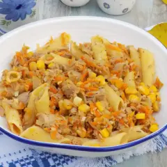 Penne z tuńczykiem, kukurydzą i startą marchewką w białej misce na niebiesko-białej serwetce.