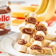 Naleśniki z nutellą i bananem na patyczkach, obok słoik nutelli i banany. Pyszna przekąska!