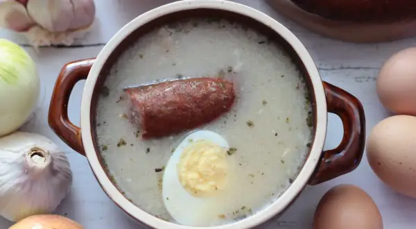 Aromaticzny zakwas na żurek w glinianym naczyniu, z kiełbasą i jajkiem. Obok czosnek, cebula i jajka.