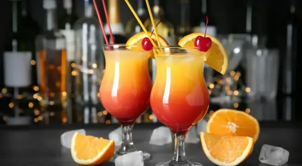 Dwa drinki Tequila Sunrise, ozdobione pomarańczą i wiśnią, stoją na barze. W tle widać butelki alkoholu i migoczące światełka. Idealne na wieczór z przyjaciółmi, jak w najlepszych lokalach Baczewski.