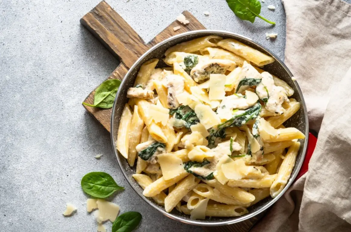 Penne arrabiata z kurczakiem i szpinakiem, posypane parmezanem, podane w patelni na drewnianej desce.