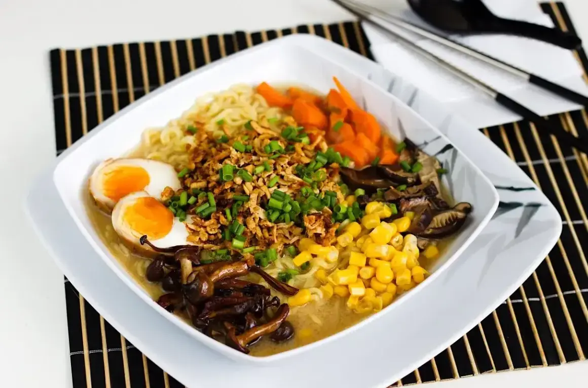 Miseczka aromatycznego tantanmen z makaronem, jajkiem na pół, kukurydzą, marchewką i grzybami shiitake, posypana szczypiorkiem.