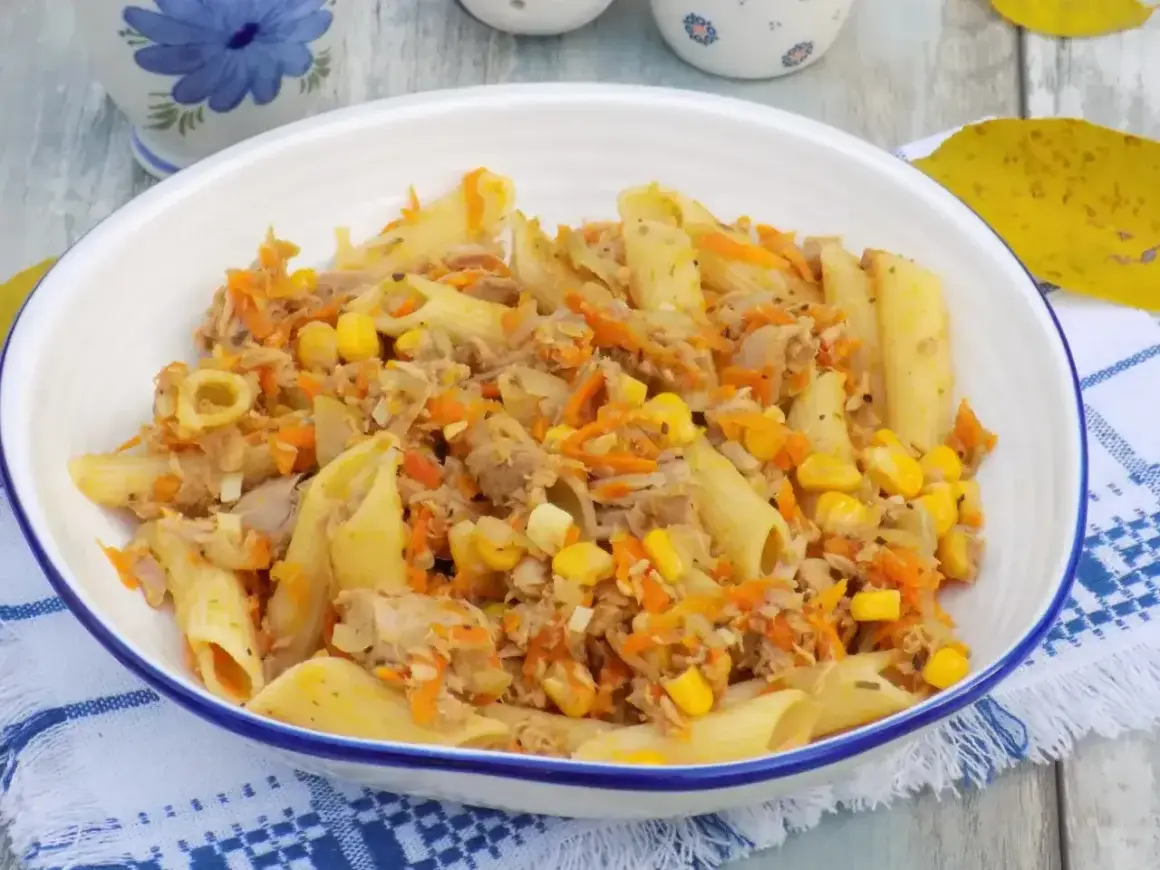 Penne z tuńczykiem, kukurydzą i startą marchewką w białej misce na niebiesko-białej serwetce.