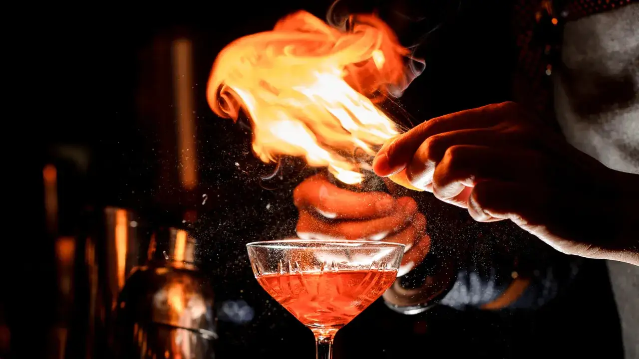Mistrz barmański wykonuje flambirowanie, tworząc efektowny płomień nad kieliszkiem z czerwonym drinkiem.