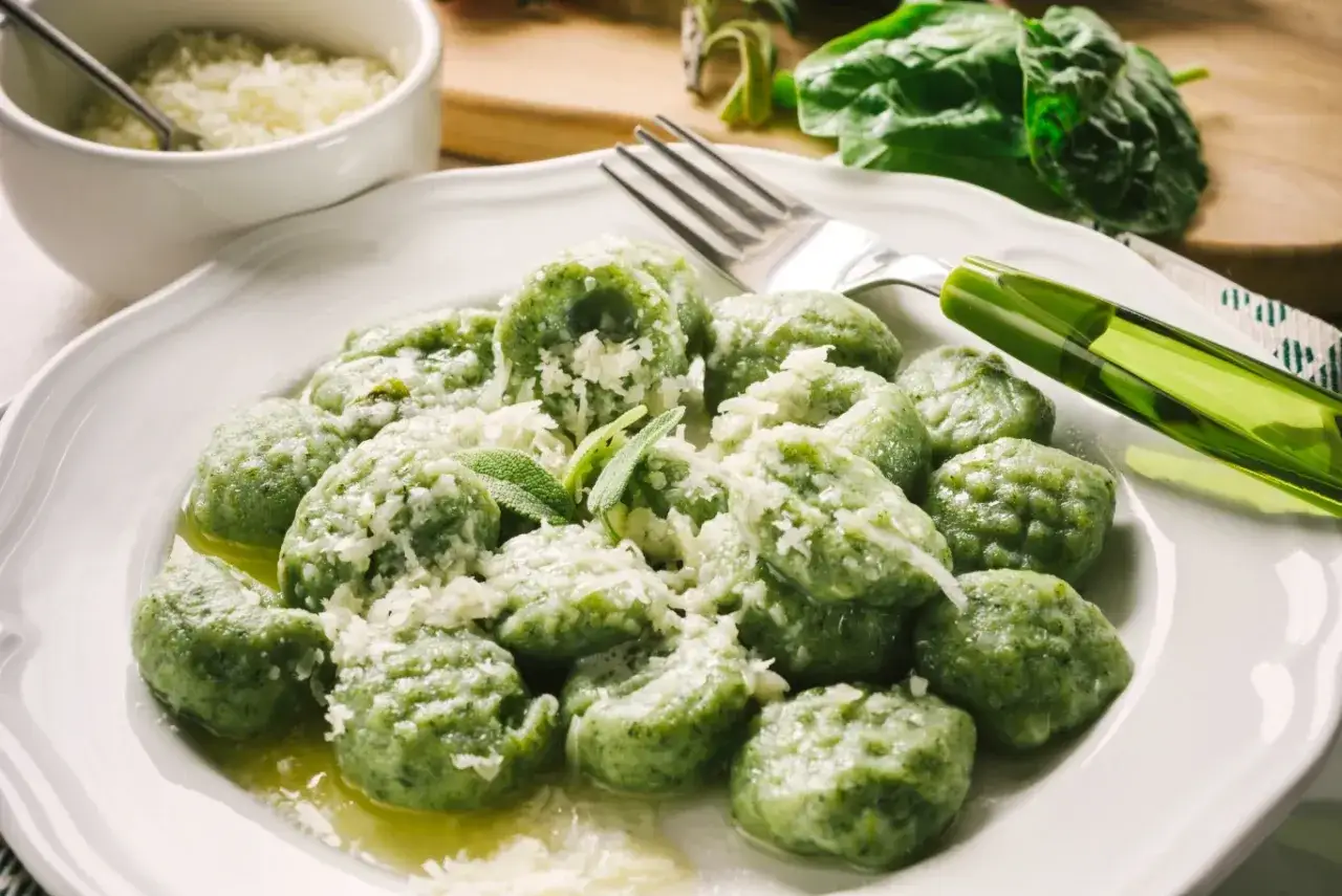 Zielone gnocchi ze szpinakiem, posypane parmezanem, podane na białym talerzu z gałązką szałwii.