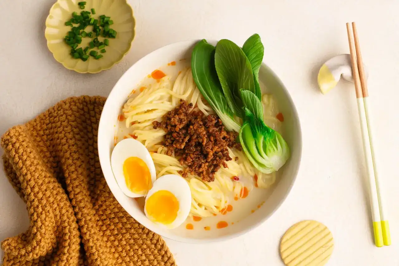 Miseczka tantanmen z mielonym mięsem, jajkiem i pak choi. Obok pałeczki i miseczka z zieloną cebulką.