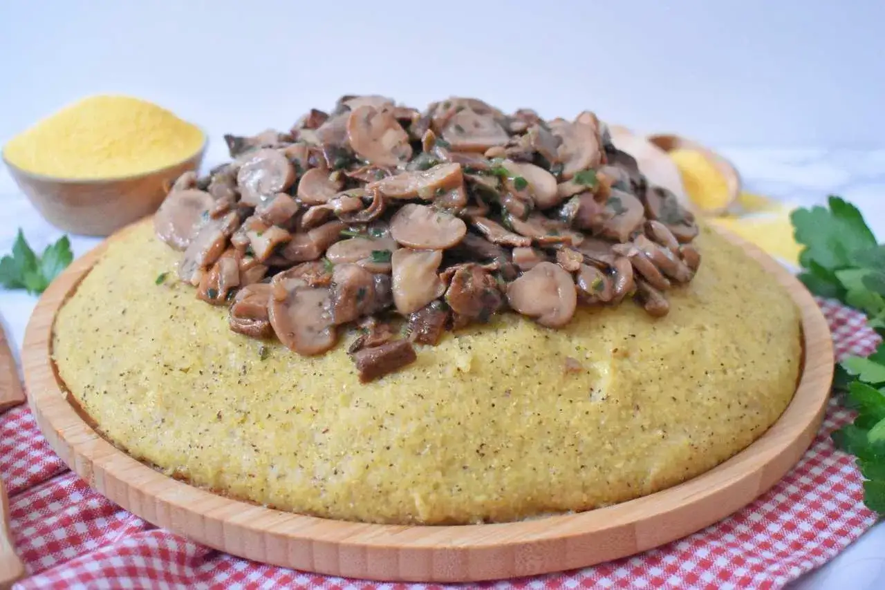 Puszysta polenta z aromatycznymi, duszonymi grzybami na drewnianej desce, ozdobiona natką pietruszki. Obok miseczka z mąką kukurydzianą.
