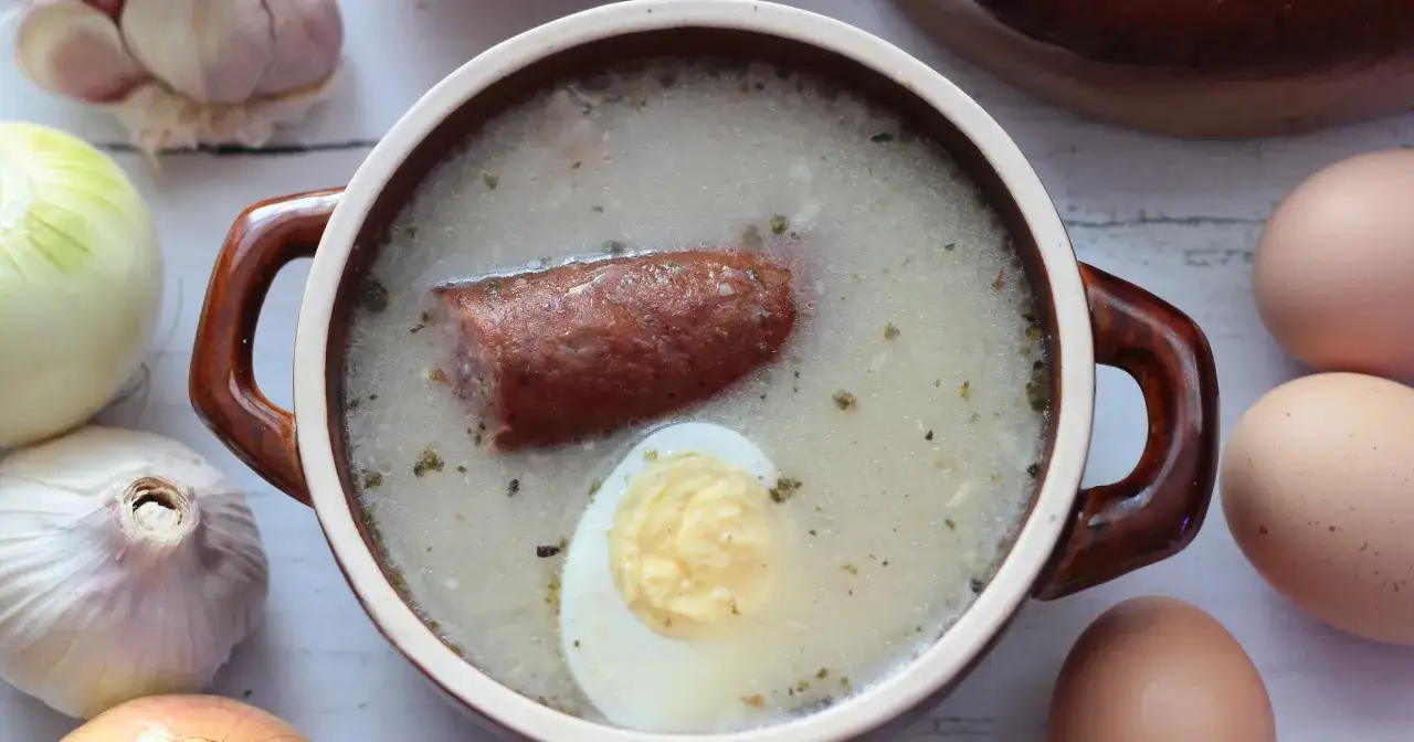 Aromaticzny zakwas na żurek w glinianym naczyniu, z kiełbasą i jajkiem. Obok czosnek, cebula i jajka.