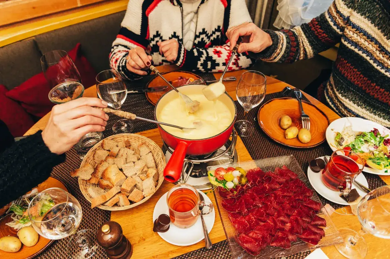 Rodzina delektuje się pysznym serem raclette, maczając chleb w roztopionym serze. Na stole widać też wędliny, ziemniaki i sałatkę.